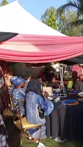 The video shows an outdoor event, possibly a wedding or celebration, taking place under several large white tents with pink and black draping. People dressed in traditional African attire are seen milling around, some seated at tables covered with black cloths. The event takes place on a grassy area, surrounded by tall trees. A musical accompaniment is audible, adding to the festive atmosphere.