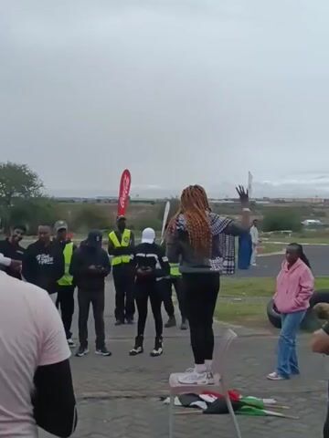 A woman is standing on a chair and addressing a group of people in an outdoor setting. The woman has long braids, is wearing black pants, a gray top with white writing on the back, and white sneakers. She is gesturing with her right arm. The people are standing and listening to her. Most of them are men. Some are wearing vests with the colors yellow and black. There are banners behind the group, and a tire lying on the ground. The environment appears to be a racetrack. The sky is overcast.