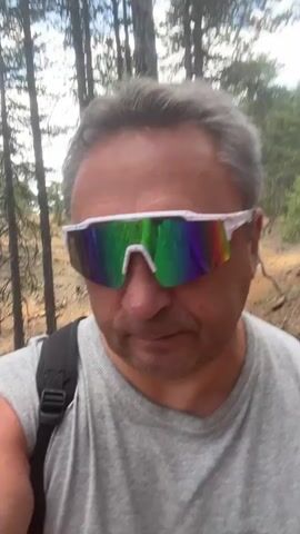 A man wearing white framed, mirrored sunglasses and a gray t-shirt is walking on a dirt path in a forest. He is looking at the camera and appears to be filming himself. The background is filled with tall pine trees and rocky terrain. The man is wearing a black backpack.