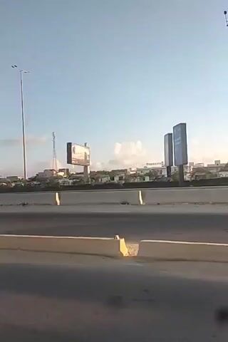 The video shows a highway scene at sunset. The sun is setting on the right side of the frame, casting a bright glare and creating long shadows. There are several streetlights and billboards visible along the side of the highway. In the background, there is a city skyline with buildings. The highway appears to be elevated, with concrete barriers separating the lanes. The sky is blue with a few scattered clouds. The video is shot from a moving vehicle, likely a car, as indicated by the perspective and motion blur.