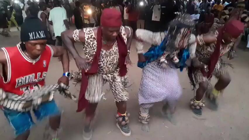 The video shows four individuals engaged in a rhythmic dance within an outdoor setting. The group is part of a larger crowd visible in the background. The lighting indicates daylight. The dancers are all adorned in colorful costumes. On the left, one dancer sports a red Chicago Bulls jersey over a blue shirt, coupled with a black "Miami" beanie. Next to him, another dancer is clad in an animal print jumpsuit, accented by a red head wrap and scarf. The third dancer is wearing a white and blue patterned fabric over his costume, which includes a matching headpiece. The last dancer is attired in a similar outfit to the second dancer, with a maroon head wrap and scarf. All dancers wear ankle bells, contributing to the rhythmic sounds. The backdrop consists of trees and a large crowd of onlookers. The ground is covered in sand.