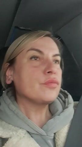 A woman with blonde hair is in a car. She is wearing a gray hoodie with a cream-colored lining. She has a stud earring in her left ear and is wearing a seatbelt. She is looking to her right and pursing her lips. Music is playing in the background. The interior of the car is visible.