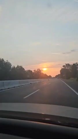 The video shows a first-person perspective of driving on a highway at sunset. The sun is visible in the distance, low on the horizon, casting a warm orange glow across the sky and reflecting on the road. Trees line the sides of the highway, and a concrete barrier separates the lanes. There are a few other vehicles visible on the road ahead. The sky is a pale blue with some wispy clouds.