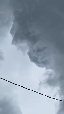 This video captures a low-angle view of a dark, stormy sky with heavy, gray clouds. A thin, black wire runs diagonally across the lower portion of the frame, obscuring a small part of the clouds below it. The clouds are dense and appear to be moving, with some areas of lighter gray and others of deep, ominous dark gray. The overall impression is one of an approaching storm. There are no people or distinct objects visible other than the wire.