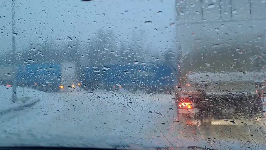 The view is from inside a car, looking through a rain-covered windshield. The focus is on the raindrops clinging to the glass, obscuring the scene outside. Beyond the windshield, there are vehicles, including a blue truck on the left and a white truck on the right, with their rear lights visible. The road appears to be covered in snow or slush. The overall environment suggests a cold, wet, and wintry day.