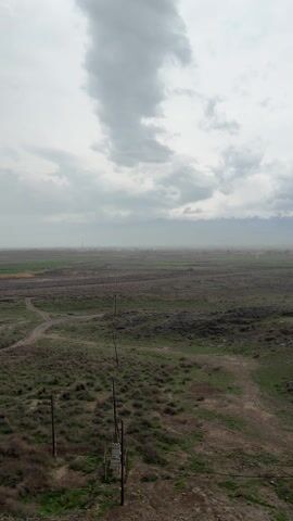 The video presents a wide, flat landscape under an overcast sky. Predominantly, the ground is covered in a mix of sparse green vegetation and brown, dirt-like patches. The landscape features multiple dirt tracks that crisscross the area.  In the foreground, a weathered wooden utility pole stands with wires extending across the view. In the background, there are hints of green fields and possibly cultivated land.  A faint suggestion of mountains can be seen on the distant horizon. The overall impression is one of a rural or agricultural setting under gloomy weather conditions.