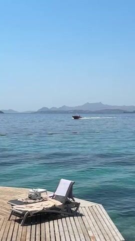 A clear blue sky is visible above a calm, blue ocean. In the foreground, a wooden deck juts out into the water, with a lounge chair and a small table. A white towel is draped over the lounge chair. In the distance, a small red boat speeds across the water, leaving a white wake behind it. Hills or mountains can be seen in the far distance on the horizon. The water is a clear turquoise color near the deck, transitioning to a deeper blue further out.