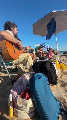 A man with a beard and sunglasses sits on a beach chair playing an acoustic guitar. He is wearing a white shirt and tan pants. There are two women sitting behind him, one in a bikini and one in a pink top. They are sitting under a blue umbrella. In the foreground, there are bags and towels on the sand. The sky is clear and blue, and the sun is shining.