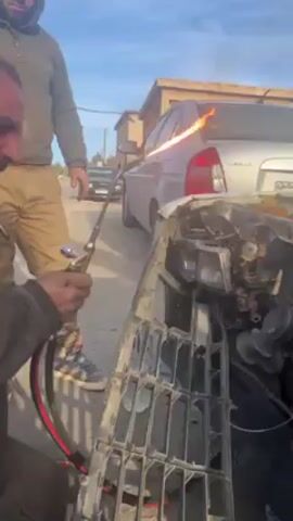 Here's a description of the video:

The video shows two men working on the front of a damaged car. One man is using a torch to heat something on the car's front end, where the grill is removed exposing the radiator and other parts. The grill is also leaning in front of the car. The second man is standing behind him observing the first man. The car is in poor condition with visible rust and damage. Another car, silver, is parked behind it. The setting appears to be an outdoor area, possibly a street or repair shop. The weather is sunny.