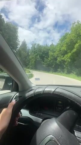 The video shows the interior of a car driving on a winding road through a forest. The camera is positioned behind the steering wheel, facing forward. A person's hand with manicured nails is visible on the steering wheel. The car is moving at a moderate speed, and the scenery outside the windows is dominated by lush green trees and a cloudy blue sky. The road appears to be paved, and there are occasional guardrails visible along the sides. The dashboard of the car is partially visible, with some warning lights illuminated. The overall impression is one of a scenic drive through a natural landscape.