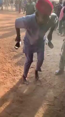 The video shows a group of people gathered in a sandy, possibly rural area. The central figure is a man wearing a light-colored, long-sleeved outfit, a red cap, and is barefoot. He is holding a pair of sandals in his hands and appears to be dancing or moving energetically. Other people are surrounding him, some are recording the scene with their phones, and others are singing and clapping. The environment appears to be an outdoor space with buildings visible in the background. The lighting suggests it is daytime.