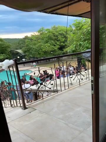 The video opens on a group of people gathered outdoors around a swimming pool, with a railing and a balcony in the foreground. The sky is cloudy and the trees are lush and green, suggesting a warm, possibly humid climate. The group appears to be a mix of adults and children. Some individuals are holding what appear to be musical instruments, possibly guitars. A person wearing a pink shirt and another wearing a white shirt are prominent within the group. The people are mostly facing the camera, with some engaged in conversation. The pool itself is visible to the left, with a tiled edge and what looks like a white decorative element. The overall impression is of a casual gathering or event taking place in a semi-tropical or resort-like setting.