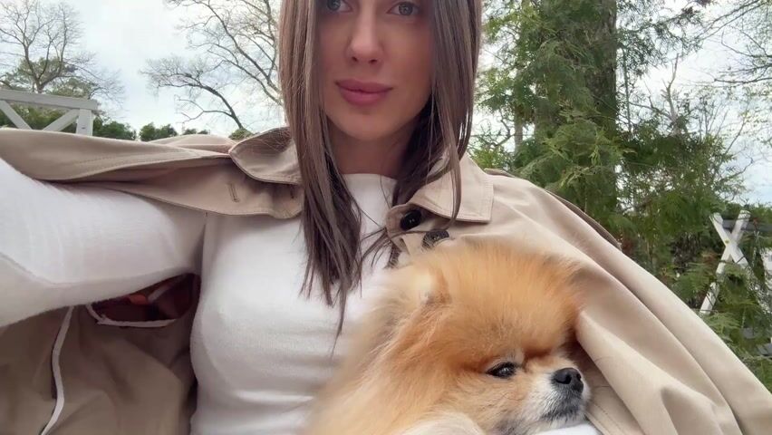 A young woman is holding a small, light brown dog, possibly a Pomeranian, in her arms while recording a selfie-style video. She is wearing a white long-sleeved shirt and a beige trench coat. The coat is draped over her shoulders and partially covers the dog. The woman is smiling slightly, looking directly at the camera. In the background, there are trees, a white wooden fence, and an overcast sky. The setting appears to be outdoors, possibly in a garden or park.