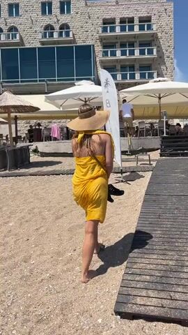A young woman with blonde hair wearing a wide-brimmed hat and a yellow sarong walks on a sandy beach past a wooden walkway. She is carrying a surfboard with a logo on it. In the background, there is a building with windows and balconies, several white umbrellas, and people sitting at tables. The sky is blue and clear.