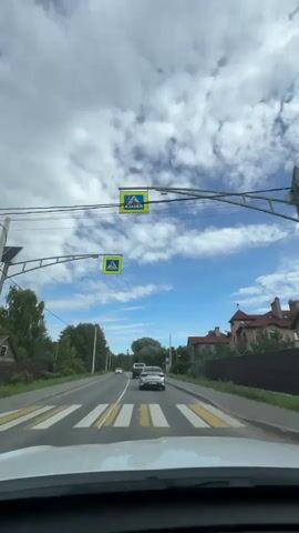 The video shows a view from a car driving on a road. The sky is blue with white clouds. On the left side of the road, there are trees and a utility pole. On the right side of the road, there are houses and a fence. There are other cars on the road ahead of the video's car. A pedestrian crossing sign is visible on a pole on the left side of the road. The video appears to be taken during the daytime.