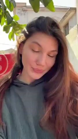 A young woman with long, reddish-brown hair stands outside in front of buildings, looking directly at the camera. She is wearing a dark green, ribbed hoodie. She smiles slightly. A hand appears and briefly adjusts her hair. There are green leaves in the upper left corner of the frame. In the background, there is a clothing rack with colorful items hanging on it, along with a two-story building with a white facade. The scene appears to be sunny.