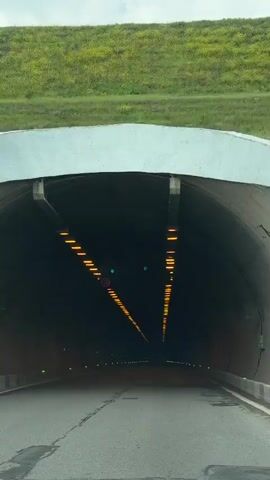 The video shows a car driving through a tunnel. The tunnel has rows of yellow lights on the ceiling and green lights along the sides of the road. There are also speed limit signs visible. A dance track plays throughout the video.