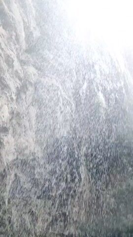 The video shows a close-up shot of a waterfall cascading down a rocky cliff face. The water appears as a misty spray, obscuring the details of the rocks beneath. The lighting suggests it is daytime, with some sunlight filtering through the mist, creating a bright, ethereal effect. The overall impression is one of natural beauty and the raw power of water.