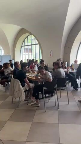 The video shows a group of young people sitting around tables in what appears to be a dining hall or common room. The room has high, arched ceilings and large windows, suggesting an older building. The floor is tiled in a checkerboard pattern of beige and brown squares.  Several individuals are visible, some seated at the tables, others standing or walking. In the foreground, two young men are seated at a table, one in a black t-shirt and the other in a white t-shirt, both appearing to be engaged in conversation or activity at the table. Other people are seated around the tables, with some interacting with each other. There are drinks and objects on the tables. A young man in a gray t-shirt is standing and looking towards the seated individuals. The lighting is bright, coming from the windows and possibly overhead sources. The overall atmosphere appears casual and social.