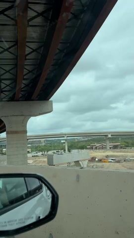 The video shows a view of Fiesta Texas amusement park in San Antonio from a moving car. The camera looks up at elevated highway overpasses. Through the concrete structures, the Six Flags park can be seen in the distance, including rollercoasters, a tall drop tower called "Fiesta Texas," and a Longhorn Steakhouse restaurant. The car is moving along a highway with concrete barriers. The sky is overcast.