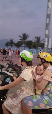 A woman with a white and green helmet is driving a black scooter with its headlight on. She has a passenger behind her wearing a yellow helmet and holding a baby in her arms. The woman driving is smiling. They are riding on a paved area lined with parked scooters on one side and a beach with palm trees and people on the other. The sky is dusky, suggesting it is either early morning or evening. There is a blue and white road sign visible on the left side of the frame.