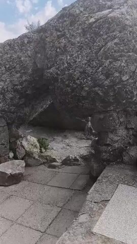 The video pans across a rocky, outdoor landscape under a cloudy sky. In the foreground, a paved area with square tiles leads towards a cluster of large boulders. One boulder appears to overhang a gap, creating a dark recess. Some smaller stones and debris are scattered around the base of the boulders. As the video moves to the right, a dense bush with green leaves and some red and white objects, possibly plastic bags or fabric, is visible growing amongst the rocks. A rudimentary set of metal steps, partially covered by vegetation, is also visible leading upwards amongst the rocks. The overall impression is one of a natural, somewhat unkempt environment with evidence of human presence, including the steps and discarded materials.