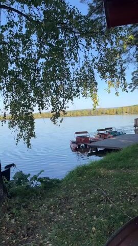 A still shot shows a calm lake with a wooden dock. Two red paddle boats are tied to the dock. The foreground is grassy with fallen leaves. The background features a treeline across the lake and a clear blue sky. A tree with green leaves hangs over the left side of the frame.