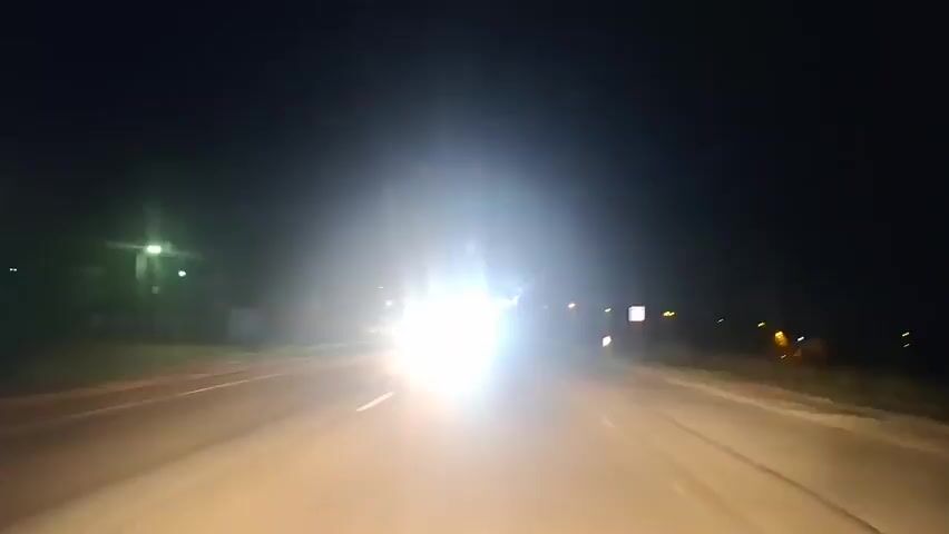 A first-person perspective video shows a view from inside a car driving at night on a road. The road has lane markings, and there are streetlights visible on the left side. Several oncoming cars with bright headlights create glare. There are also some signs and distant lights visible on the right side of the road. The sky is dark.