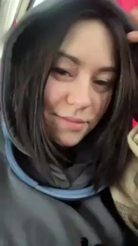 A woman with dark hair wearing a gray hooded sweatshirt and a blue neck pillow is sitting on a bus with red curtains in the background. She looks at the camera and smiles slightly.