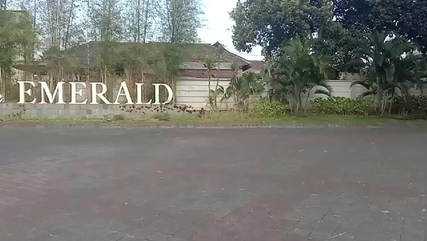A wide shot shows a sign that reads "The EMERALD" in large white letters. The sign is positioned in front of a wall and bamboo trees. To the left of the sign, there is a billboard with images and text. The road in front of the sign is paved with dark asphalt and has a brick border. The surrounding environment appears to be a residential or commercial area with buildings and vegetation visible in the background. The lighting suggests it is daytime, possibly early morning or late afternoon due to the soft light. No people are visible in the frame.