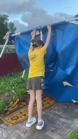 A person, seen from behind, stands with their arms raised, holding onto the edge of a large, collapsed blue tarp. They are wearing a bright yellow t-shirt with text on the back, dark grey shorts with white ribbon accents, white socks, and white platform shoes. Their hair is pulled back in a bun. The tarp appears to be part of a structure, possibly a canopy or tent, which has fallen over. The person is standing on a patterned rug laid out on the ground, which is covered with grass and dirt. In the background, there is a wooden fence, greenery, and trees. The sky above is cloudy, suggesting it might be late afternoon or early evening. The overall scene appears to be an outdoor setting, likely a backyard or a rural area. The text on the t-shirt is in Russian. The transcribed text in the video is "Ты хочешь вообще его убирать?" which translates to "Do you want to remove it at all?". This suggests a conversation about dismantling or removing the collapsed structure.