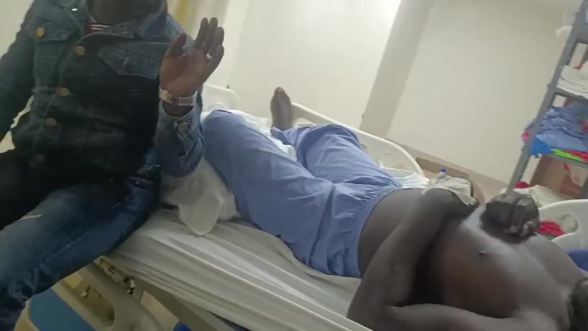 In a medical setting, a man in blue scrubs lies in a hospital bed. He is shirtless, and his eyes are closed. Another man, wearing a denim jacket and jeans, sits on the edge of the bed next to him. The man in the denim jacket appears to be speaking, gesturing with his hands. In the background, there is a metal shelving unit holding folded linens and medical supplies. The room is brightly lit.