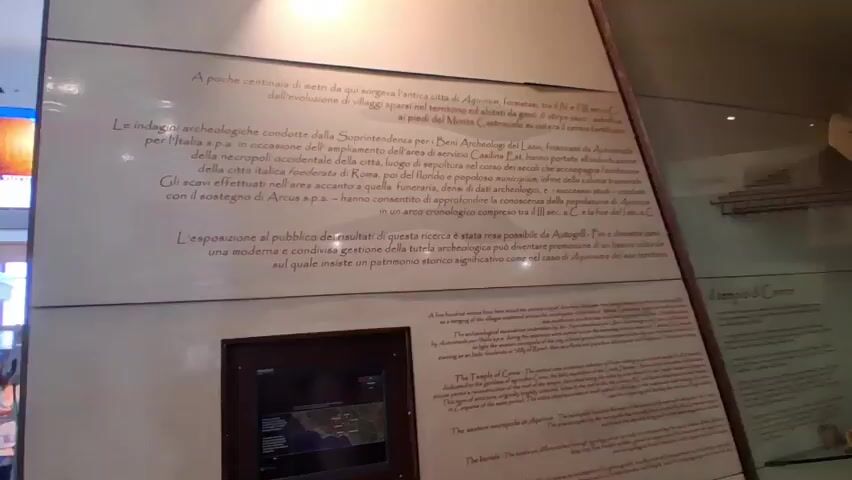A video showcases a wall with text panels in both Italian and English. The Italian text, written in a light brown font, is displayed prominently on a large, white panel. Below this, a smaller section of the wall features English text in a similar font. The overall environment appears to be an indoor exhibition space. In the foreground, a dark-framed monitor is visible, displaying a map or informational graphic. To the far left of the frame, a person in a white top is partially visible, facing away from the camera. The camera is angled upwards, capturing the lower portion of the wall display.