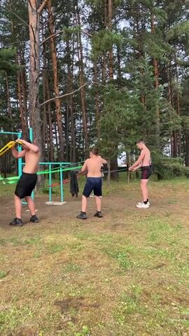 Four boys are exercising with resistance bands in a forest clearing. A metal jungle gym with turquoise bars is in the center of the frame. The boy on the far left, wearing black shorts and a white t-shirt, is holding a red resistance band. The boy next to him, wearing black shorts and a black t-shirt, is holding a green resistance band. The boy in the middle, wearing blue shorts and a white t-shirt, is holding a green resistance band. The boy on the right, wearing black shorts and a white t-shirt, is holding a yellow resistance band. The boys are all facing the jungle gym and appear to be engaged in some form of training or game. The ground is covered in grass and dirt. Tall pine trees are visible in the background. The sky is overcast.