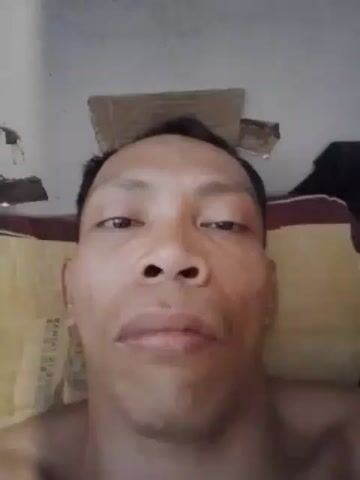 The video displays a close-up, selfie-style shot of a person's face, likely filmed from a low angle. The individual has dark hair and appears to be looking directly at the camera with a neutral expression. The background is out of focus, but hints of a pillow and a textured wall are visible. The lighting is somewhat harsh, casting shadows on the face. The overall impression is of a raw, unfiltered moment captured by the individual themselves.