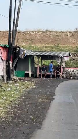 The video shows a roadside scene with a simple shelter. Three individuals are seated or standing within or near the shelter. The shelter appears to be constructed of basic materials, including a corrugated metal roof and possibly wooden or metal supports. Laundry is hanging on a line or pole to the left of the shelter. The ground around the shelter is unpaved and appears dusty or muddy. A paved road curves into the foreground. In the background, a dry, grassy embankment rises. Electrical wires are visible overhead, supported by poles.