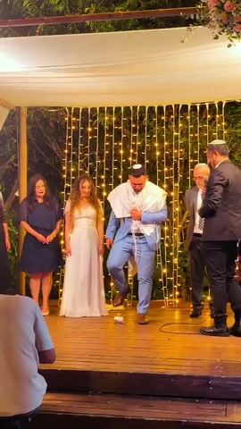 A wedding ceremony is taking place outdoors under a canopy decorated with white fabric and string lights. A bride in a white dress and a groom in a light blue suit stand on a wooden platform. The groom is wearing a tallit over his shoulders. Several people are observing the ceremony. On the left, a woman in a dark blue dress stands with her hands clasped. Next to her, the bride is smiling. On the right, a man in a suit and tie, possibly an officiant, stands with his hands clasped. Another man in a suit is visible to his right. In the foreground, a person wearing a light brown t-shirt is visible from the back, holding a phone. The overall atmosphere appears festive, with guests clapping and cheering.