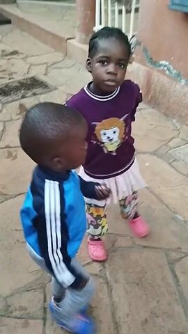 Two young children are standing on a tiled patio. A girl with braided hair is wearing a purple sweatshirt with a picture on it, a white skirt, patterned leggings, and pink Crocs. She has her hands on her hips and is looking towards the other child. The other child is a young boy wearing a blue jacket with white stripes, grey pants, and blue Crocs. He turns to look at the girl and salutes. The patio is made of various-sized tiles in shades of tan and brown. Behind the children is a wall with a metal box attached to it. A white metal railing can be seen in the background.