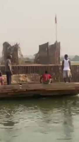 The video shows people and a white van on a large wooden boat. The boat is moving across a body of water. There is a small structure on the side with a red flag attached. People stand on the boat and the van is being driven on. On the far bank, an excavator is visible. The sky is overcast.