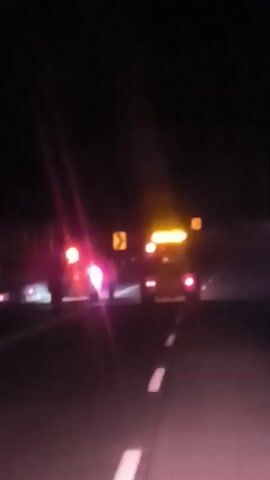 The video captures a night-time scene on a highway. The primary subjects are several vehicles, including what appears to be a truck and a smaller vehicle, possibly a car or van, in the distance. The truck has multiple red taillights and yellow overhead lights. The smaller vehicle also displays red taillights. The road is paved and marked with white lane lines. The overall setting is dark, suggesting it is nighttime. There are also yellow warning signs visible, indicating curves in the road. The video is blurry, likely due to the low light conditions and movement. There is no visible text in the media. The activity appears to be vehicles traveling on a highway at night. The emotions conveyed are neutral, given the lack of clear human subjects or discernible facial expressions. The location is noted as unknown but contextually Indonesia.