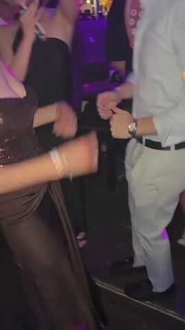 The video shows the interior of a crowded nightclub with people dancing to music. The lighting is purple and blue, and there are pink balloons floating near the ceiling. A DJ booth is visible in the background with a DJ performing.  The crowd is mixed, some dressed casually, and others in more formal attire. Some people are holding drinks, and others are recording the scene with their phones. One person wears a large calculator prop on their back. The overall atmosphere is energetic and lively.