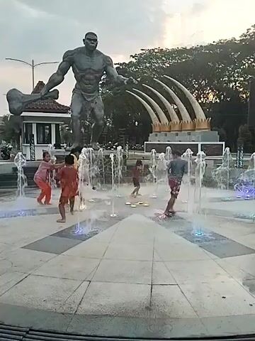 Several children play in a circular interactive fountain with colorful lights in the foreground, with one child in red playing with the water in the center, and a group of three children in yellow and red clothes running through the water in the background. Behind them, a large gray statue of a muscular ape-like figure holding a bone in its right hand stands on a pedestal, facing left. To the right of the ape statue, another monument features large white curved tusks. Lush green trees and buildings are visible further in the background under an overcast sky.