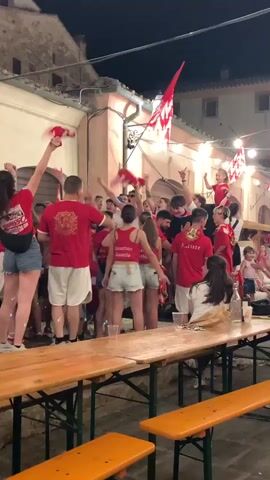 The video shows a group of people, mostly wearing red shirts and some with red and white checkered accents, gathered in what appears to be a narrow street or alley at night. String lights are hung overhead. Many individuals are raising their arms, some holding red cloths or flags, suggesting a celebration or event. Several people are seen dancing and interacting with each other. In the foreground, there are wooden picnic tables and benches. Some individuals are on tables or being carried on shoulders, indicating a lively and enthusiastic atmosphere. The buildings in the background are stone or stucco, illuminated by the lights and the ambient glow.