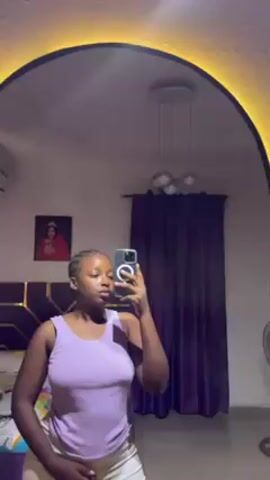 A young woman is taking a selfie in a mirror. She is wearing a light purple tank top and light-colored shorts. The mirror is framed with a light. The room behind her includes a bed with a gold headboard and a portrait hanging above the bed. Purple curtains hang on a window behind her, and a modern light fixture hangs from the ceiling. There is also an air conditioner visible in the corner of the room. The woman first poses with her lips pursed and then lowers her hand to her hip while still holding the phone.