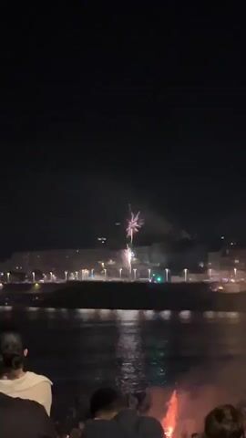 The video captures a nighttime scene with a view of a waterfront and a city skyline. In the foreground, the backs of several people are visible, facing away from the camera and towards the water and cityscape. Some of these individuals appear to be holding a fire or sparkler, which emits a reddish glow.

Across the water, a city is illuminated by numerous lights from buildings and streetlamps, suggesting it's a populated urban area. The water itself reflects these lights, creating shimmering patterns on its surface.

The main event of the video is a fireworks display. A single, large firework bursts in the dark sky, emitting a bright pinkish-purple light and sparks. Smaller fireworks or the remnants of previous ones are also visible, creating streaks of light and smoke. The overall atmosphere appears festive, likely a celebration or event drawing a crowd to the waterfront. The time of day is clearly night, and the weather appears clear, allowing for a good view of the fireworks. There is no visible text in the video.