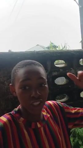 A child with dark skin and short black hair, wearing a horizontally striped shirt in red, orange, yellow, green, blue, and purple, is in the foreground. They are looking at the camera and speaking, with their mouth open in some shots. The child is standing outdoors in front of a weathered, textured wall with circular openings. Behind the wall, there is a corrugated metal roof and lush green foliage. In the background, there are two solar-powered lights mounted on poles. The lighting is overcast.