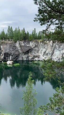 The video shows a natural landscape with a large body of water surrounded by steep, rocky cliffs. The water is a deep green color and reflects the surrounding environment. Trees, including pine and deciduous varieties, grow along the top of the cliffs and in the foreground. The sky is overcast. There are several people visible along the edge of the cliff on the far side of the water, appearing to be observing the scene. A single white rock or boulder is partially submerged in the water near the cliff face. The overall impression is one of a serene, natural quarry or gorge.