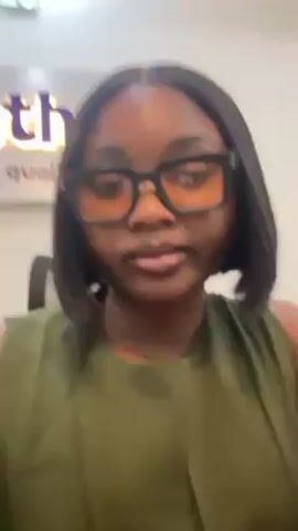 A woman with short dark hair and glasses is looking directly at the camera. She is wearing a sleeveless green top. The background is white with a sign on the wall partially visible. The sign is purple and includes the word "health". The woman's mouth is slightly open.