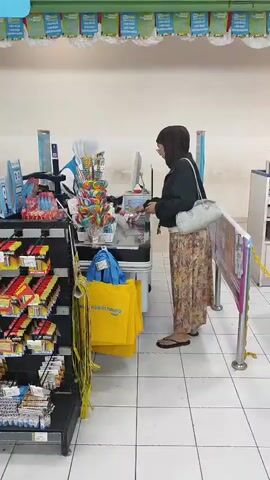 A woman wearing a hijab and a floral skirt stands at a checkout counter in a store. She is interacting with a cashier who is partially obscured by the counter. To the left of the counter, there are shelves stocked with various products. In front of the counter, there are several yellow reusable shopping bags hanging. The floor is tiled, and the walls are a light color, with green trim at the top.