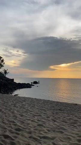 The video shows a beach at sunset. The sky is a mix of gray and white clouds, with a sliver of orange sun peeking through. The sea is calm and reflects the orange sunlight. There are a few rocky outcrops near the shore, and a person is standing on one of them. The beach is sandy and there are some tracks in the sand. As the video progresses, the sky becomes more blue and the sun disappears. Two boats are visible in the distance. An island appears on the horizon.